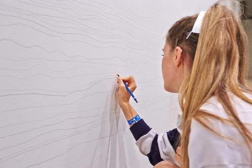 A student carefully draws her line on the wall, making sure not to touch the lines on either side. She is also trying to apply a consistent pressure and often rotates the pencil to keep the line a consistent thickness and hue.